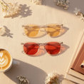 Load image into Gallery viewer, Two pairs of sunglasses with yellow and red lenses on a light surface with flowers and a cup of coffee.