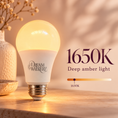 Load image into Gallery viewer, A lit Dream Walker 1650K deep amber LED light bulb with a white frosted body and metallic screw base, shown on a wooden surface with decorative dried flowers and text reading '1650K Deep amber light' and a 1650K slider graphic.

